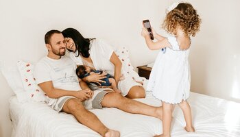 A family of four on the bed with a small girl taking a picture of the parents and baby