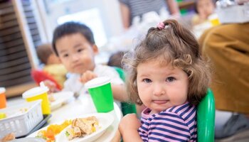 A toddler looking at the camera with a plateful of food in front of her and another toddler in the background